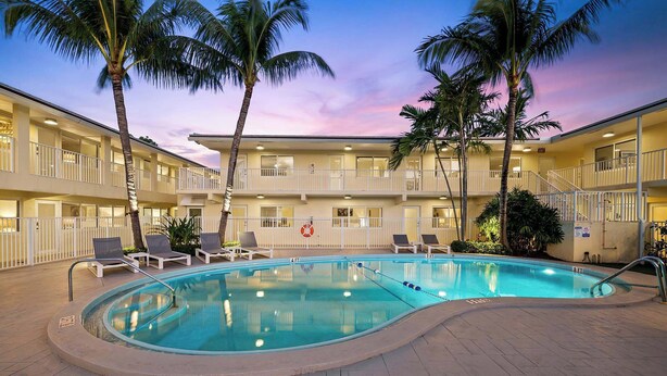 A swimming pool with lounge chairs and palm trees, surrounded by a multi-story building with balconies. - Bermuda Isles Villas - King #29 (Lauderdale-by-the-Sea)