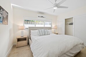 A bedroom with a bed, two bedside tables with lamps, a ceiling fan, an air conditioning unit, a window with blinds, and a wardrobe with a mirror.