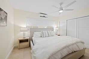 A bedroom with a bed, bedside tables, a ceiling fan, an air conditioning unit, and a window with blinds.