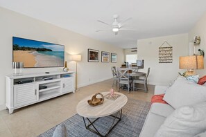 A living room with a flat-screen TV, a white sofa, a coffee table, and a dining area with chairs and a table.