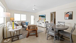 A modern living room with a sofa, a coffee table, a dining table with chairs, a lamp, and a painting on the wall. - Bermuda Isles Villas - King #32 (Lauderdale-by-the-Sea)