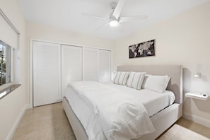 Un dormitorio con cama, ventilador de techo, ventana y estante en la pared.
