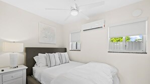 Un dormitorio con cama, mesita de noche, lámpara, ventilador de techo, unidad de aire acondicionado, ventana con persianas y un espejo fijado a la pared.