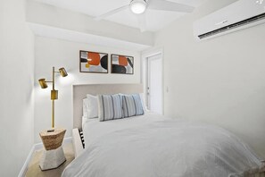 A bedroom with a bed, a ceiling fan, a wall-mounted air conditioner, and abstract art on the wall.