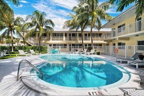 A swimming pool with lounge chairs, palm trees, and a multi-story building with balconies.