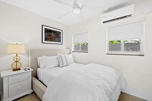A bedroom with a bed, nightstand, lamp, air conditioning unit, and a window with a view.