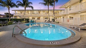Une piscine entourée de chaises longues et de palmiers, le tout bordé d’un immeuble doté de nombreux balcons.