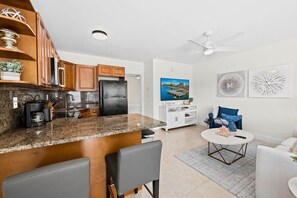 A modern kitchen with granite countertops, wooden cabinets, and a black refrigerator. There is a dining area with a round table and chairs, a white sofa, and a ceiling fan.