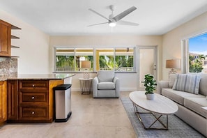 Un vaste espace de vie comprenant une cuisine, un ventilateur de plafond, un canapé, une table basse et une vue sur les palmiers, visible à travers de grandes fenêtres.
