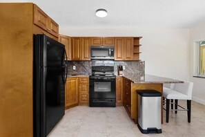 A kitchen with wooden cabinets, a black refrigerator, a microwave, an oven, and a trash bin. - Bermuda Isles Villas - Queen #17 (Lauderdale-by-the-Sea)