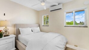 Ein Schlafzimmer mit Bett, Nachttisch, Lampe, Deckenventilator, Klimaanlage und Fenstern mit Blick ins Grüne.