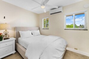 Ein Schlafzimmer mit Bett, Nachttisch, Lampe, Deckenventilator, Klimaanlage und Fenstern mit Blick ins Grüne.