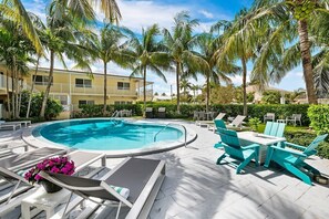 A pool area with lounge chairs and a table, surrounded by palm trees and a building in the background.