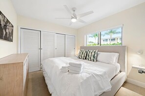 A bedroom with a bed, white curtains, a ceiling fan, a window with a view, and a wooden dresser.