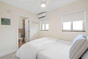 A bedroom with a bed, a ceiling fan, an air conditioning unit, a window with blinds, and a door leading to a bathroom.