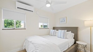 Ein Schlafzimmer mit einem Bett, Deckenventilator, Klimaanlage, Nachttischlampe und einem Fenster mit Blick auf Grünfläche.