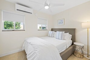 Ein Schlafzimmer mit einem Bett, Deckenventilator, Klimaanlage, Nachttischlampe und einem Fenster mit Blick auf Grünfläche.