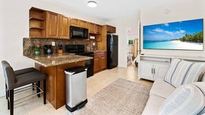 Ett modernt kök med träskåp, en inbyggd mikrovågsugn och ett kylskåp. Det finns en matplats med en stol och en soffa. En stor TV är monterad på väggen och visar en bild av en strand.