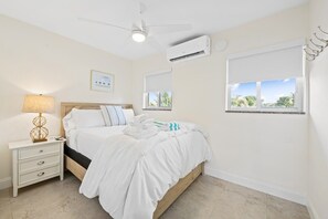 A bedroom with a bed, nightstand, lamp, ceiling fan, air conditioning unit, and a window with a view.