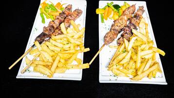 Dos platos rectangulares blancos con brochetas de carne y papas fritas.