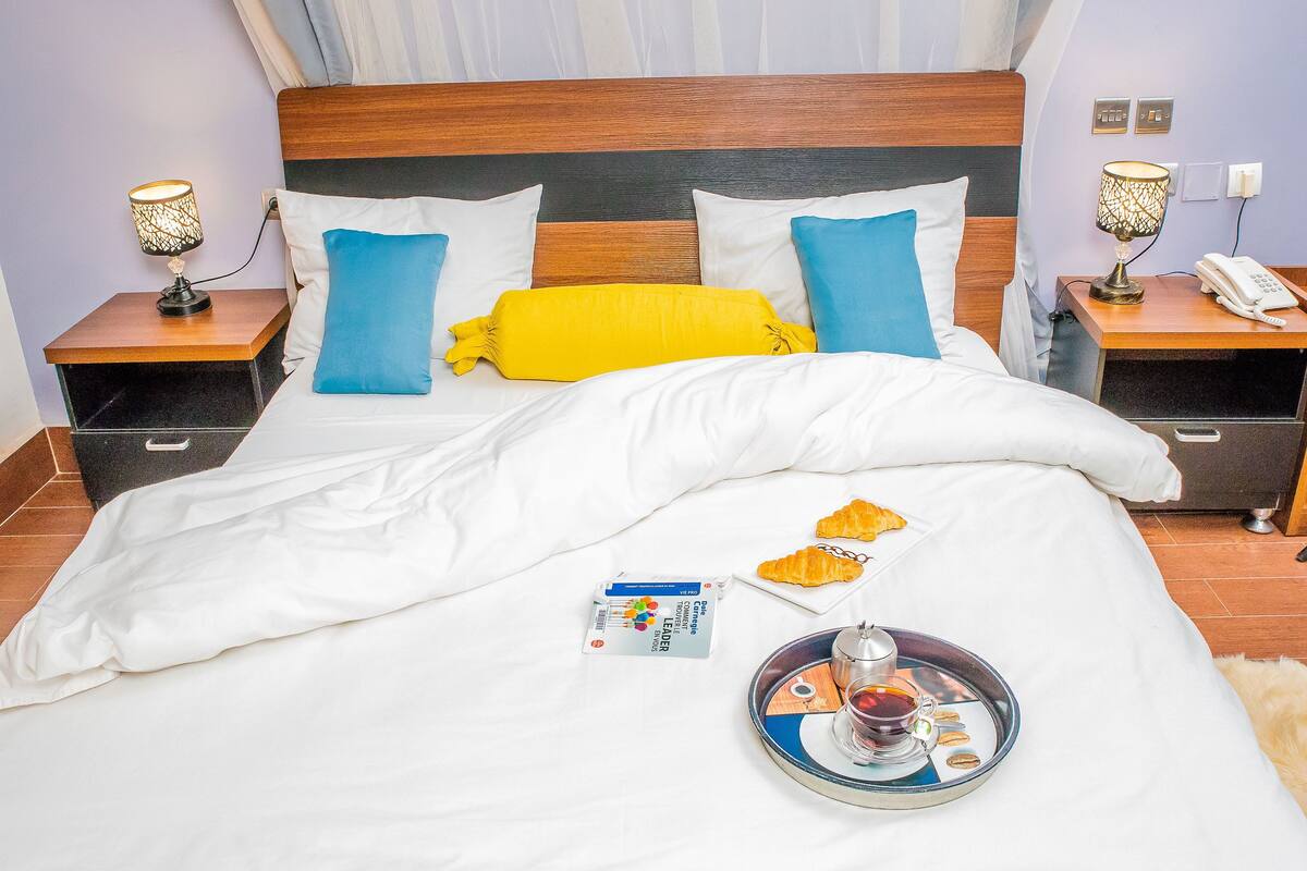 A neatly made bed with white bedding, blue and yellow pillows, and a tray with a croissant and a cup of tea.
