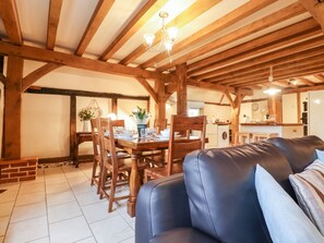 A rustic interior with exposed wooden beams, a dining area with wooden chairs and a table, a leather sofa, and a kitchen area in the background.