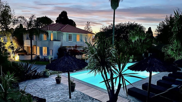 A pool area with a stone deck, palm trees, and a large house in the background.