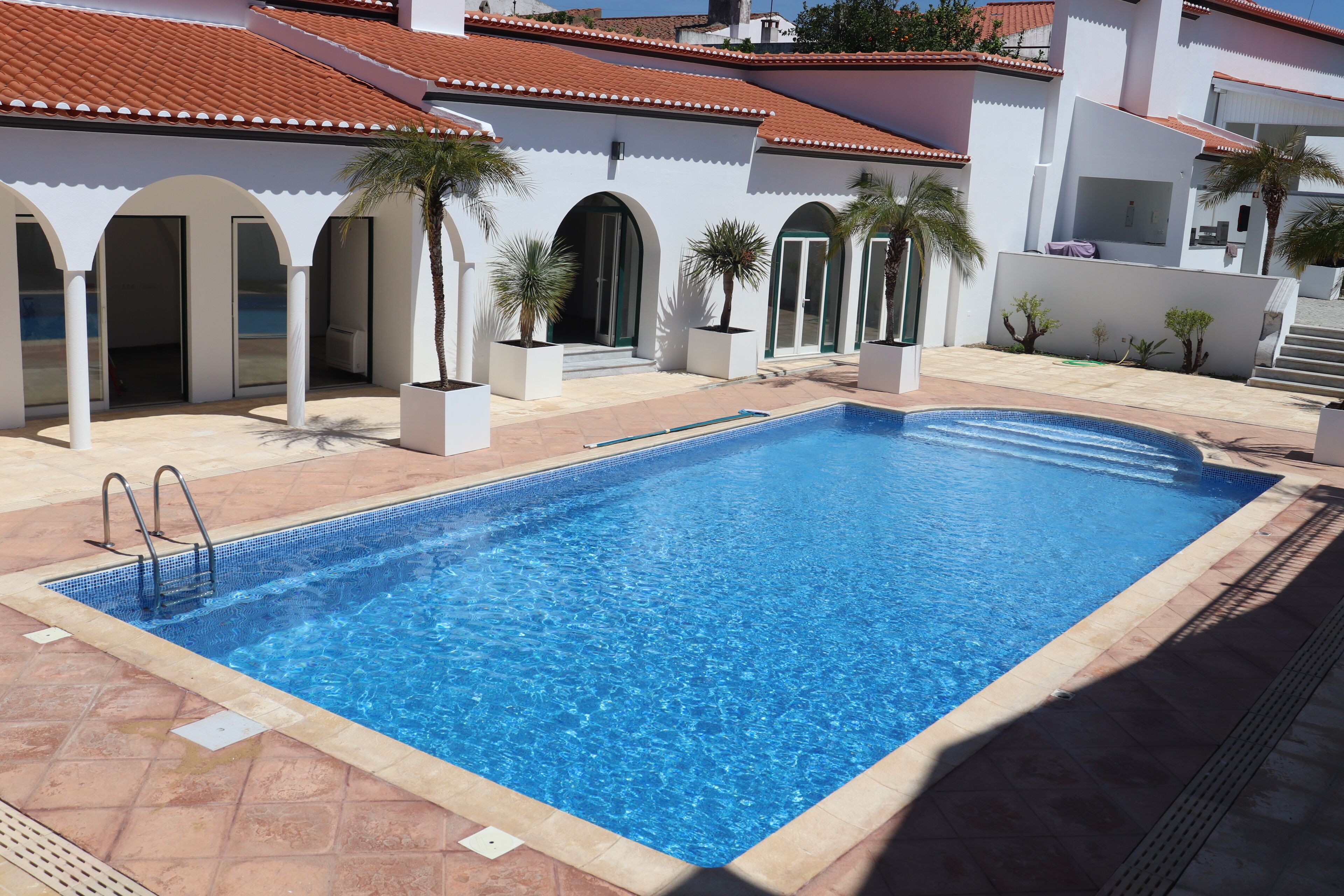 A rectangular swimming pool with clear blue water, surrounded by a tiled deck and white walls with arched doorways.