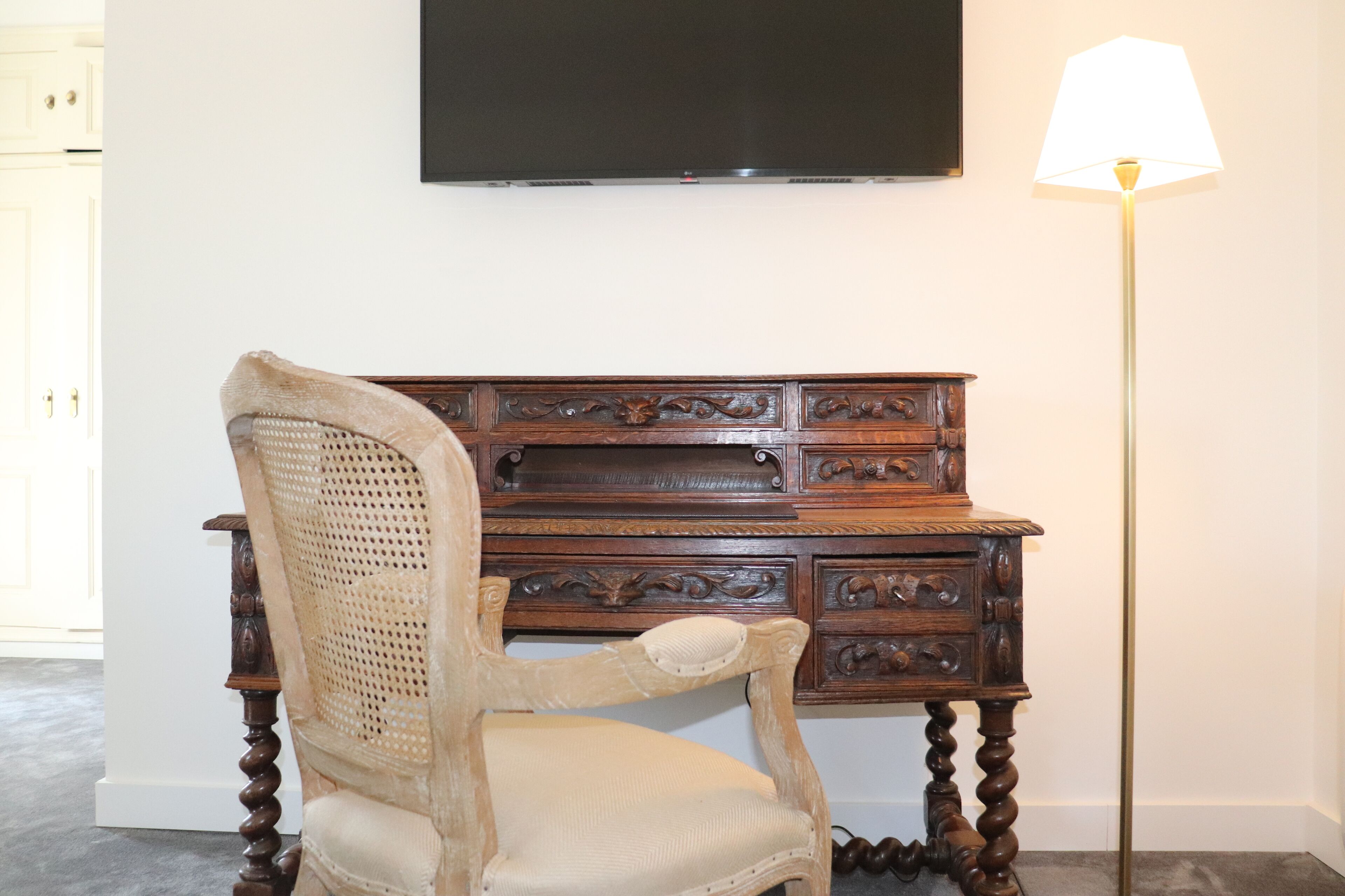 A wooden desk with intricate carvings, a wicker chair, and a floor lamp.