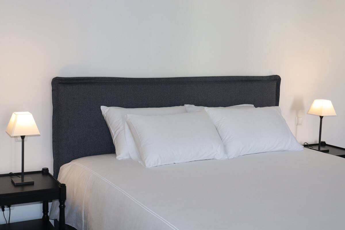 Un lit bien fait, avec une tête de lit grise, deux tables de chevet avec des lampes et un mur blanc sobre.