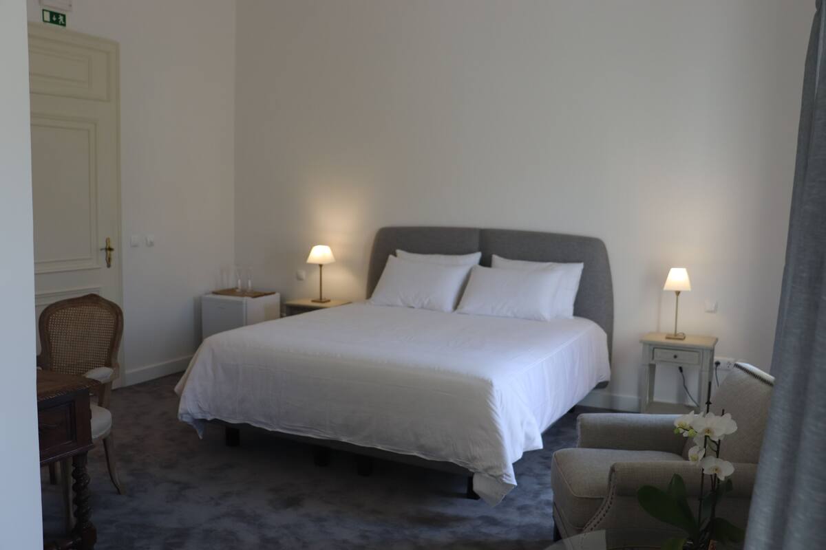 Une chambre d’hôtel bien aménagée, avec un grand lit, deux tables de chevet avec des lampes, une chaise et une petite table sur laquelle est posé un vase de fleurs.