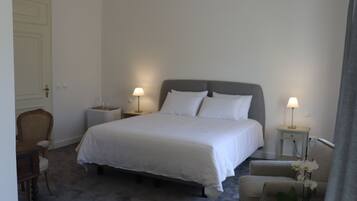 Habitación de hotel ordenada con una cama grande, dos mesitas de noche con lámparas, una silla y una pequeña mesa con un florero.