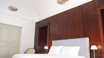 Quarto de hotel com uma cama grande, painéis de madeira nas paredes, teto com molduras decorativas e uma porta que dá para outro quarto.