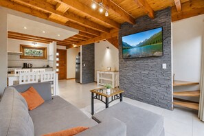 A modern living room with a grey sofa, wooden ceiling beams, a stone accent wall, a flat-screen TV, and a kitchen in the background.