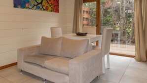 A room with a beige sofa, a small table, and a chair. There is a colorful abstract painting on the wall and a view of trees through a glass door. - Los AMANCAYS (Villa La Angostura)