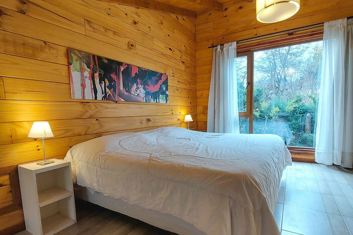 Une chambre à coucher dans une cabane en bois, avec un lit, deux lampes de chevet, un tableau au mur et une fenêtre avec des rideaux.