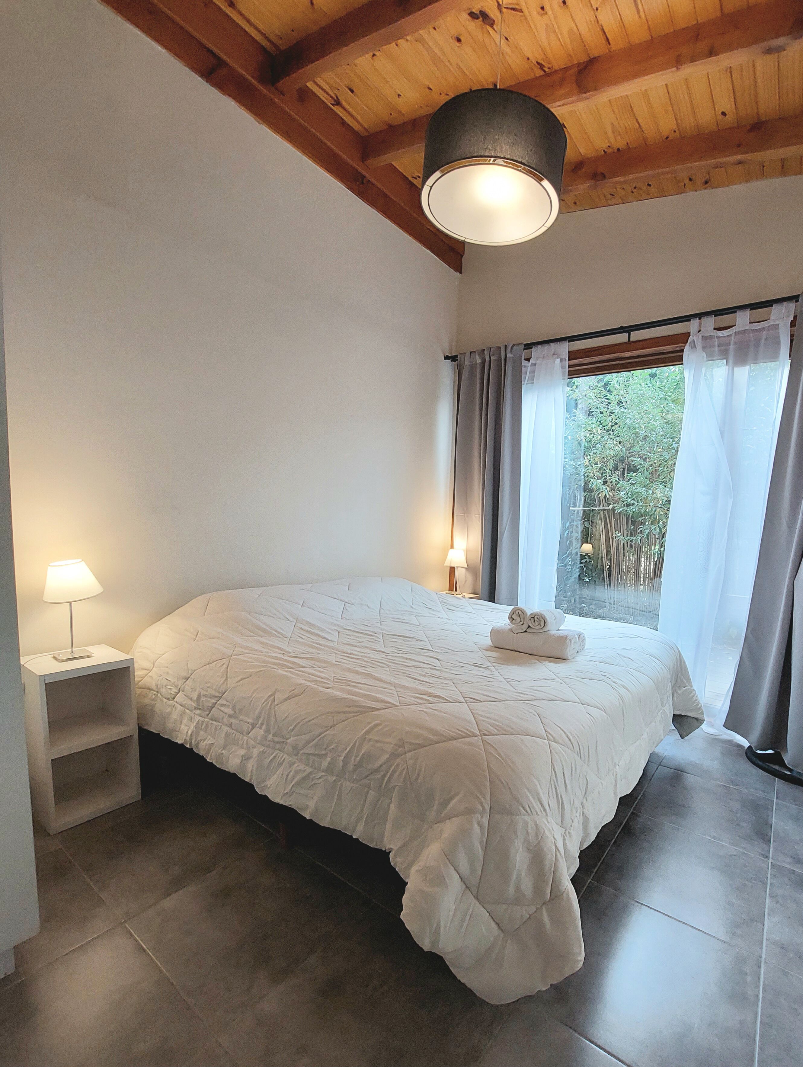 A bedroom with a large bed, a bedside table with a lamp, a ceiling light, and a window with curtains.
