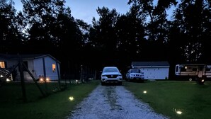 A gravel driveway leading to a white car parked on the side of a road, with a trailer and a small building on the left. - Lake House (Beechgrove)