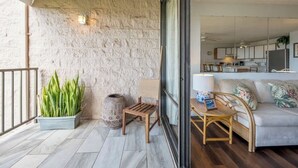 A balcony with a stone wall, a potted plant, a wooden chair, and a sofa with cushions. - Ocean view 1 bedroom, AC, walk to shops, dining, & beaches (Kihei)