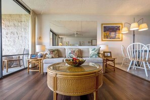 Un salon avec une table basse à plateau en verre, un canapé agrémenté de coussins à motifs floraux, un coin repas avec des chaises blanches et un grand miroir au mur.
