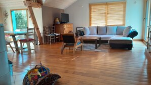 Ein gemütliches Wohnzimmer mit Holzboden, einer Couch, einem Couchtisch und einem Fernseher. Ein Fenster mit Jalousien, ein Esstisch mit Stühlen und ein Korb auf dem Boden sind ebenfalls vorhanden.