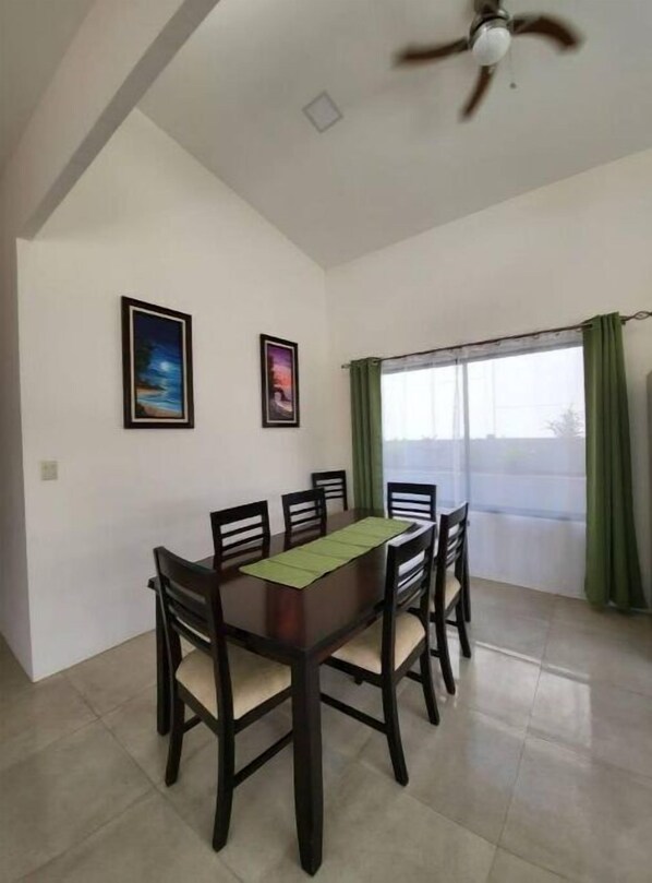 A dining area with a rectangular table, six chairs, two framed pictures on the wall, a ceiling fan, and a large window with green curtains.
