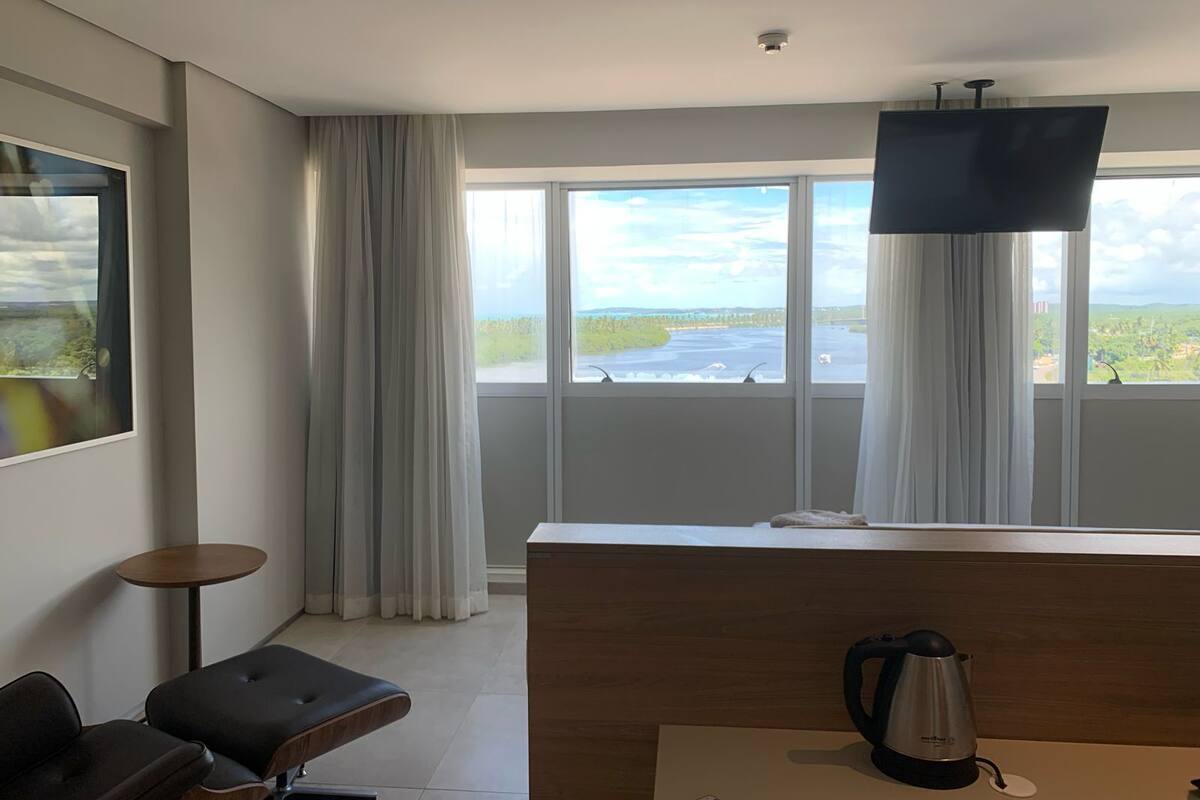 A modern hotel room with a large window, a flat-screen TV, a coffee maker, and a small round table.