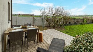 Une terrasse avec une table dressée pour deux personnes, entourée de chaises en osier, donnant sur un espace verdoyant avec un arbre et offrant une vue sur un terrain de sport.