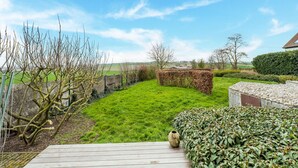 Une terrasse en bois mène à un jardin verdoyant agrémenté d’une haie, d’un mur de pierres et d’un ciel d’un bleu éclatant.