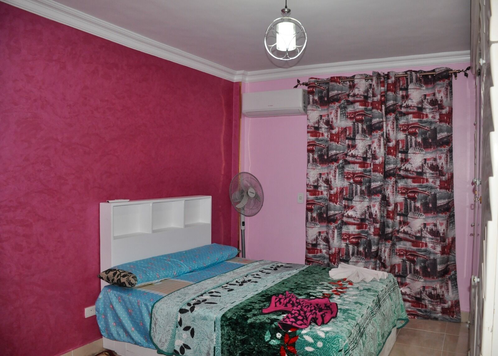 Un dormitorio con paredes rosas, un cabecero blanco, una cama con varias mantas, un ventilador de pie y una cortina estampada.