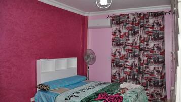 Un dormitorio con paredes rosas, un cabecero blanco, una cama con varias mantas, un ventilador de pie y una cortina estampada.