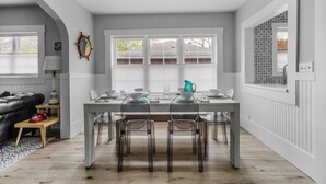 Un coin repas avec une table blanche, des chaises transparentes et une fenêtre avec des stores.