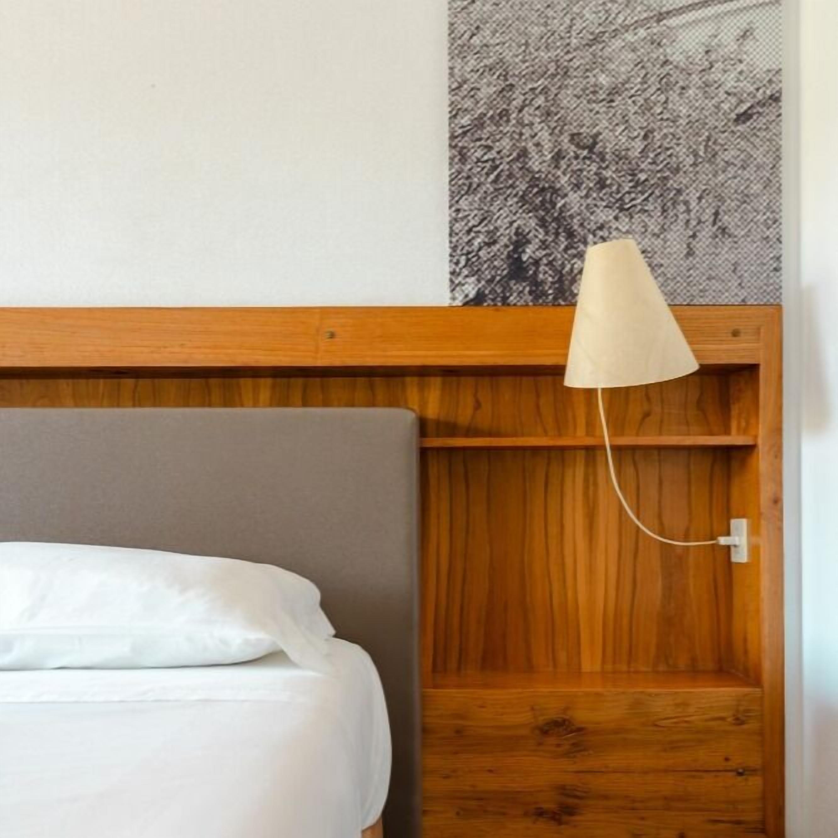 A hotel room with a wooden headboard, a bed with white bedding, and a wall-mounted lamp.