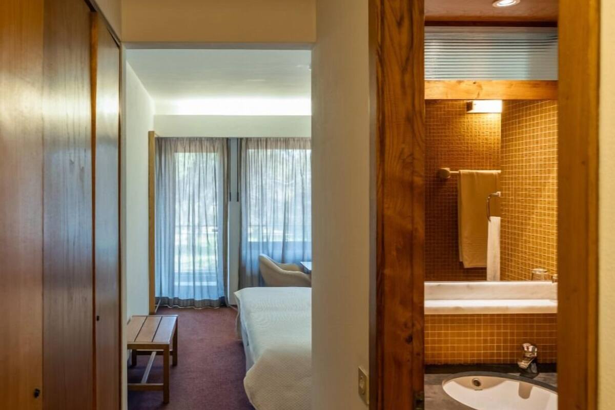 A hotel room with a bed, a small table, a bathroom with a sink and mirror, and a view through a door.